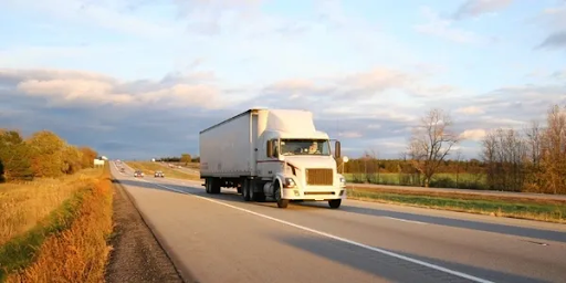 White semi-truck hauling a trailer on a rural highway through autumn fields under a cloudy sky.